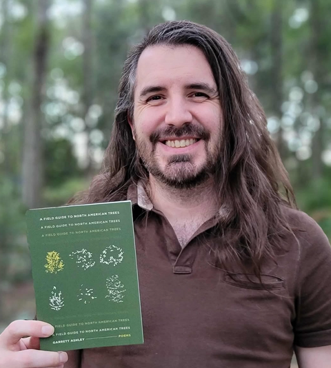 Man holding a green book with tree illustrations in a forest setting